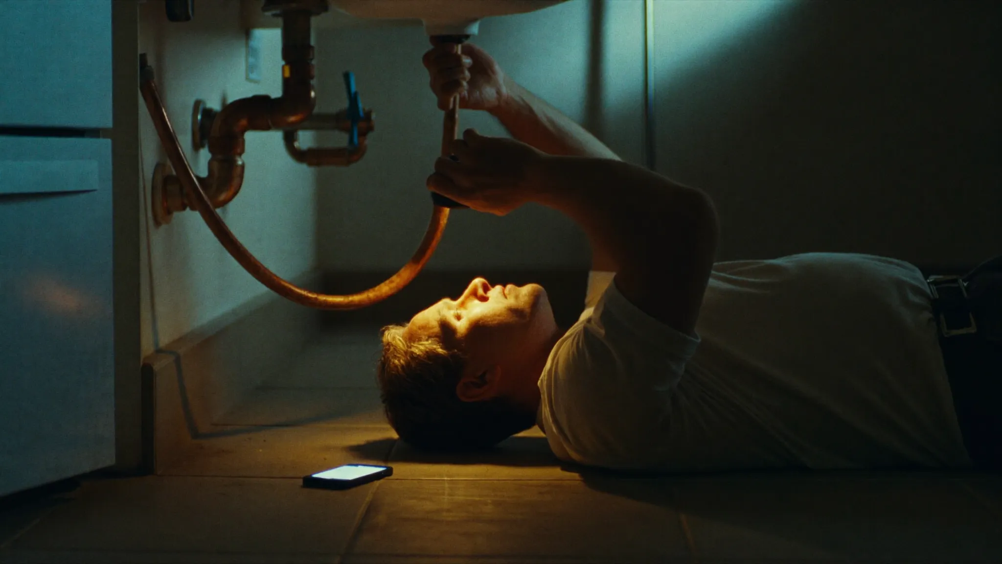 A tradesperson working under a kitchen sink by torchlight while his phone rings beside him